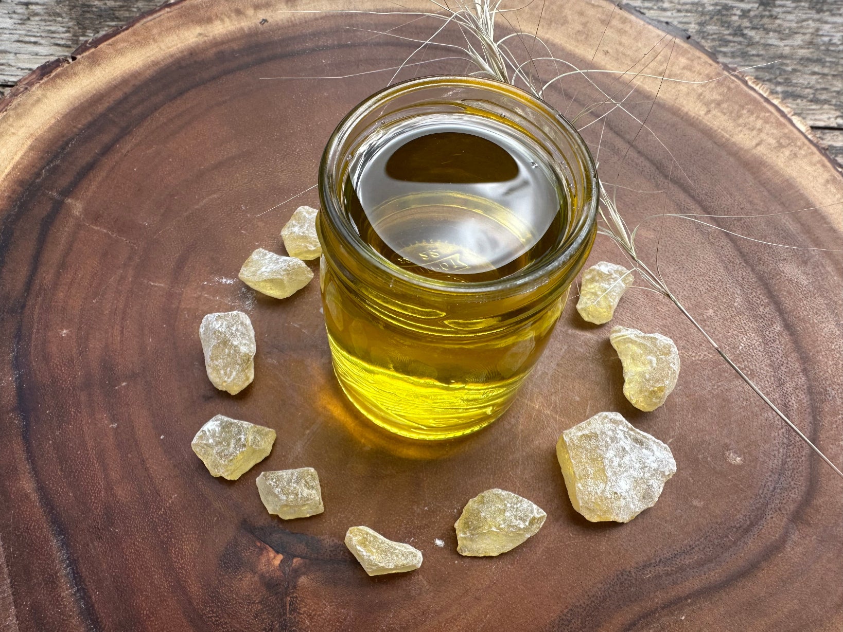 Clear glass full of golden jojoba oil is on wooden cutting board. Also on it there are some pine resin chunks and a blade of dried grass